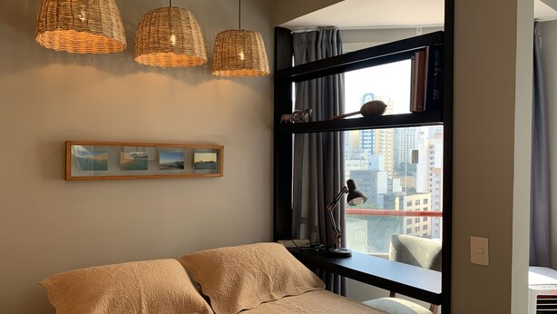 A bedroom with a bed, three hanging wicker lamps, a framed picture, and a view of a cityscape through a large window. - Akile B. Ribeiro (São Paulo)