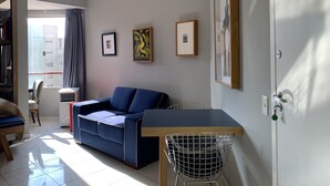 A modern living room with a blue sofa, a wooden table, and a metal chair. There are framed pictures on the wall and a window with curtains. - Akile B. Ribeiro (São Paulo)