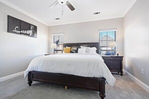 Une chambre avec un lit en bois foncé, une couvre-lit blanche, deux tables de chevet avec des lampes, un ventilateur de plafond et une grande fenêtre avec des stores.