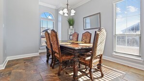 A dining room with a large wooden table, six upholstered chairs, a chandelier, and large windows with blinds. - Just minutes from LSU, SU & Downtown Baton Rouge! (Addis)
