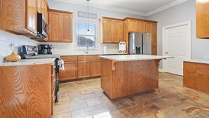A kitchen with wooden cabinets, a central island, a refrigerator, and a window with blinds. - Just minutes from LSU, SU & Downtown Baton Rouge! (Addis)