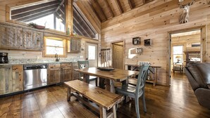 A rustic cabin interior with a wooden dining table, benches, and a kitchen area with a dishwasher and cabinets. - NEW! Private Mountain Cabin in Gated Community w/ Mtn Views, Hot Tub & Game Room (Morganton)