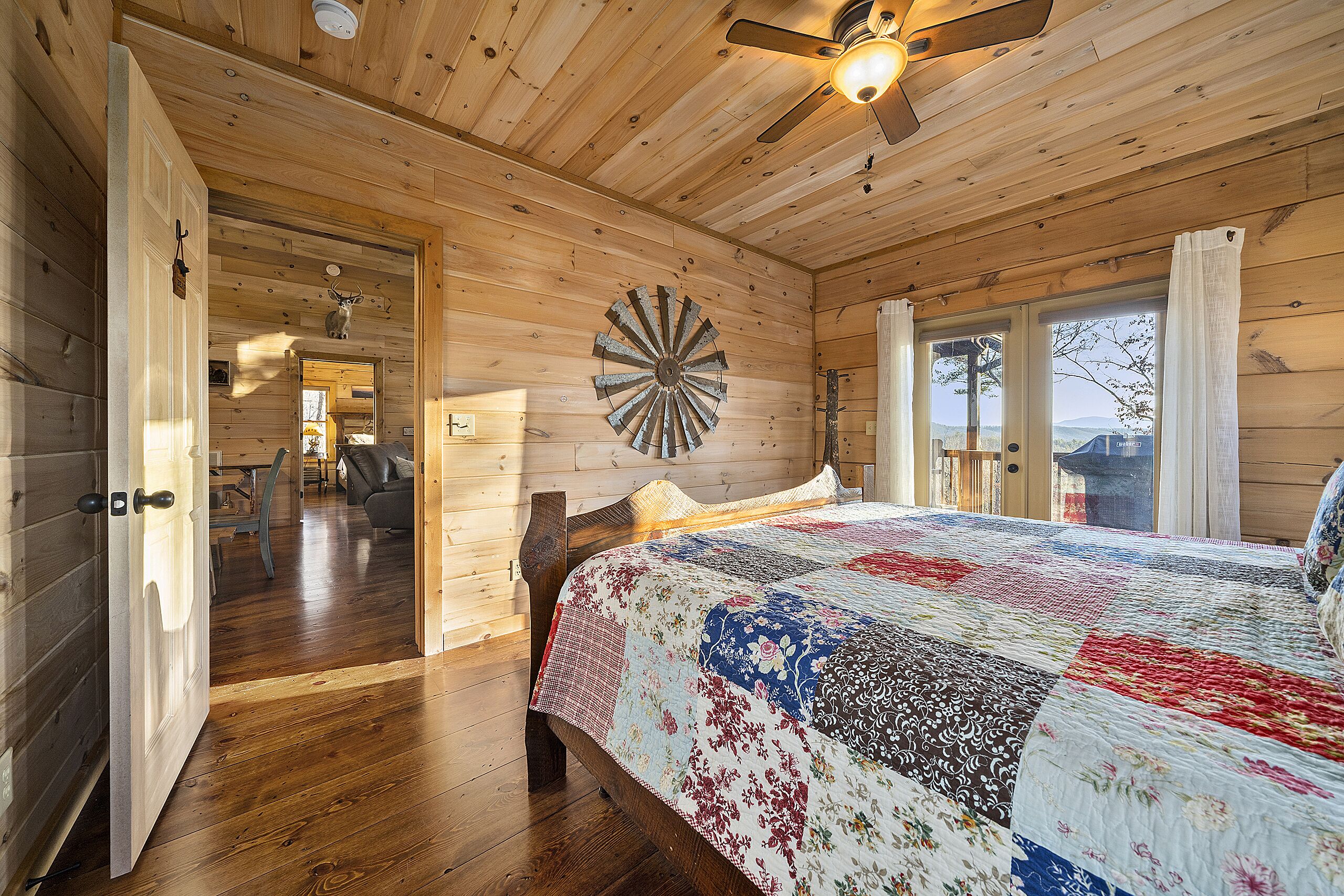 A wooden cabin bedroom with a bed, a ceiling fan, a rustic wall clock, and a large window.
