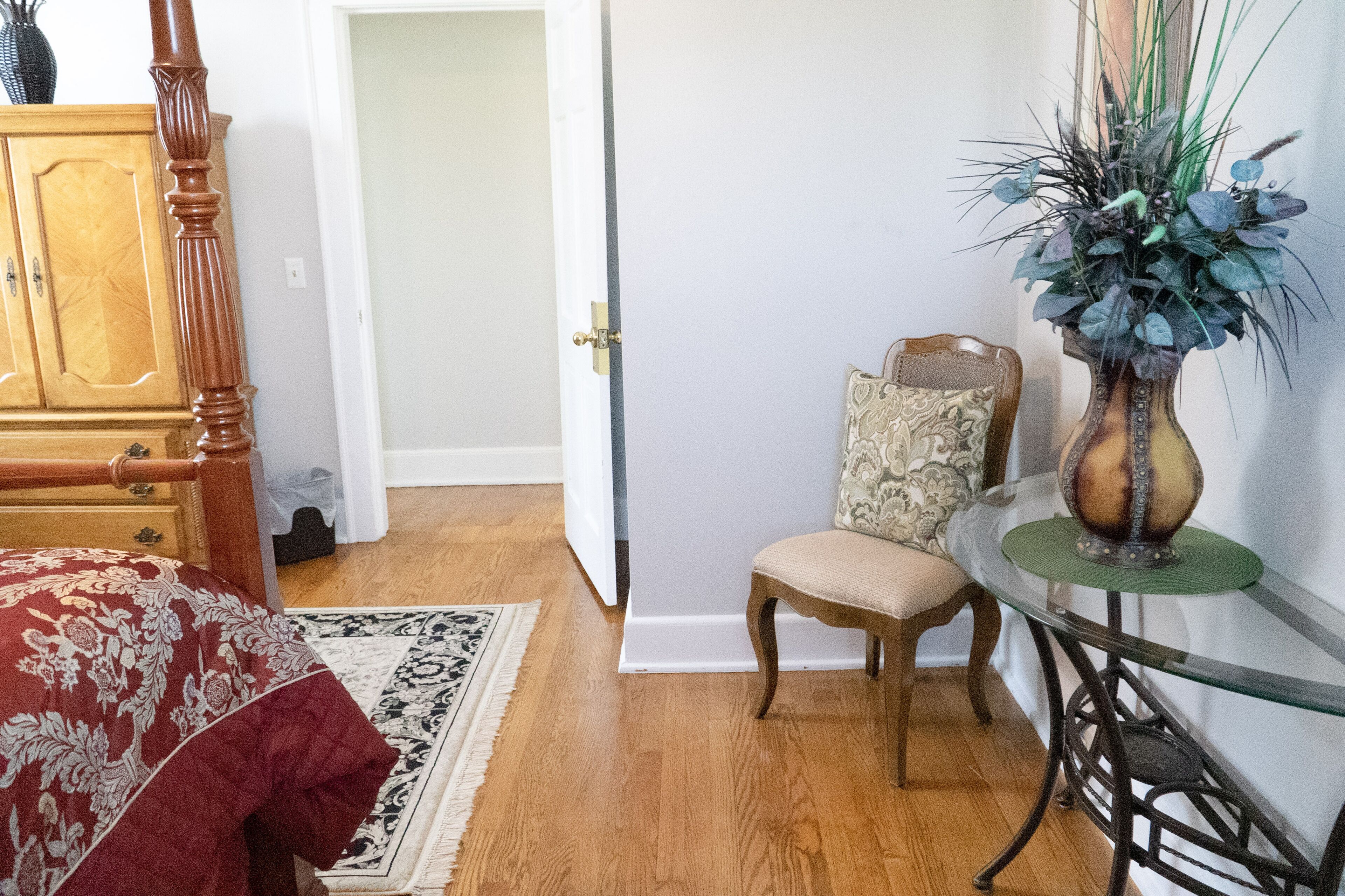 A bedroom with a wooden bed, a chair, a glass table, and a vase with plants.