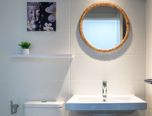 A bathroom with a white sink, a toilet, a woven mirror frame, a small potted plant, and a wall-mounted picture of a Buddha statue.