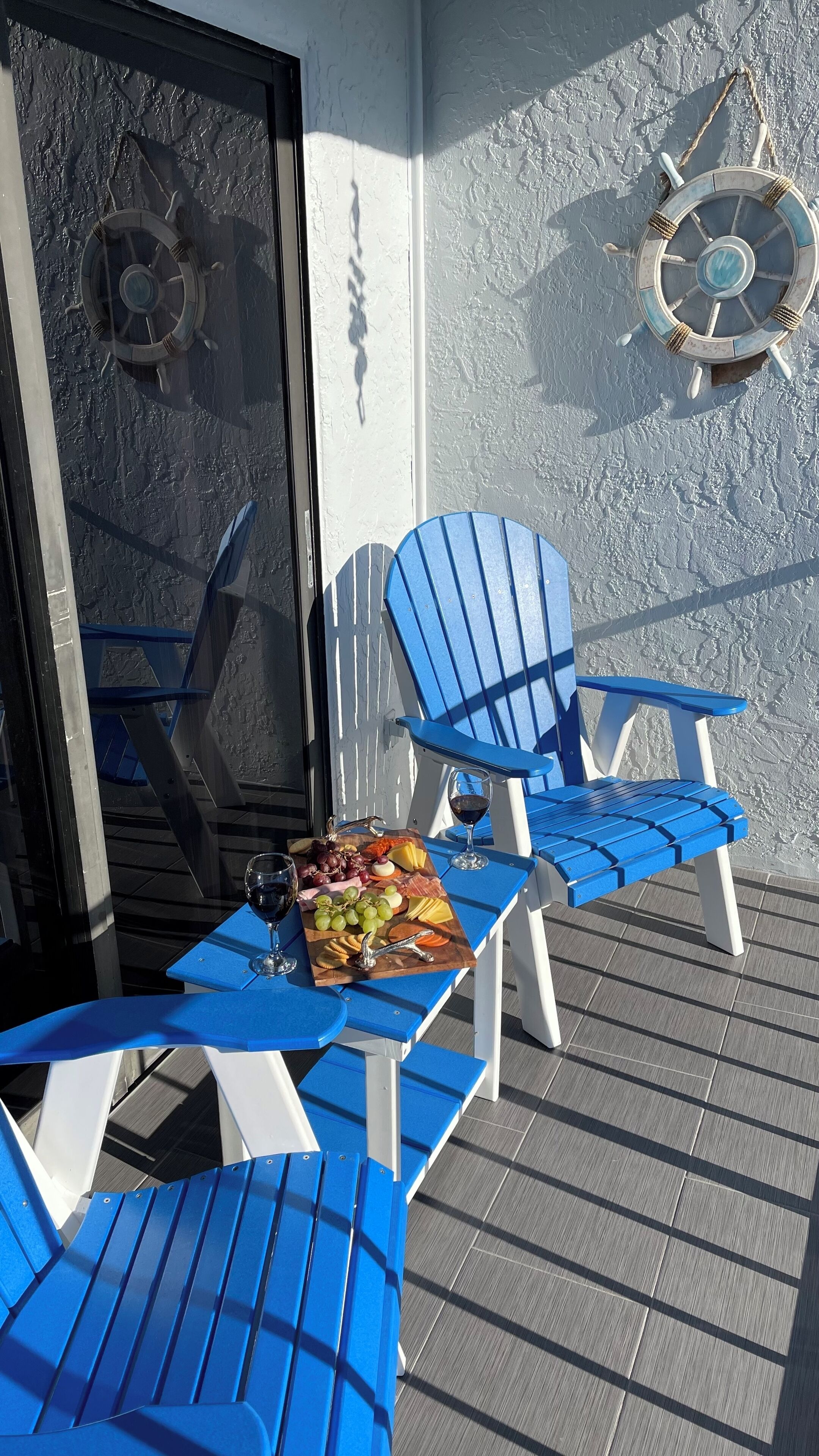 Un balcon avec des fauteuils Adirondack bleus, une table avec une bouteille de vin et une planche de fromages et de raisins.