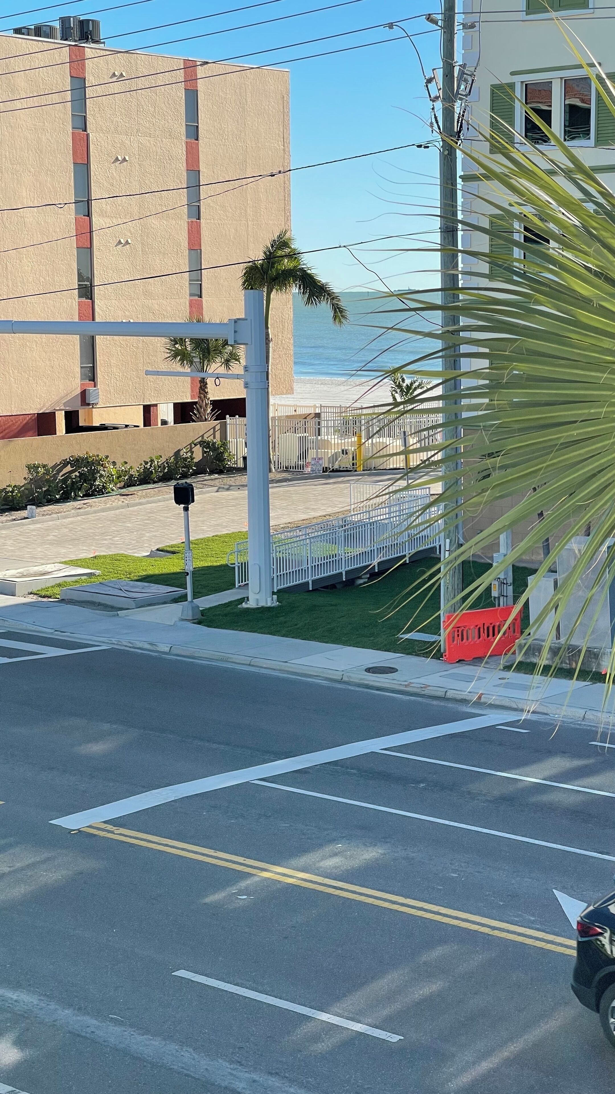 A street view with a building, a palm tree, and a beach in the background.