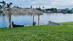 Un belvédère au toit de chaume, situé au bord d’un lac paisible, entouré d’une végétation luxuriante et de palmiers.