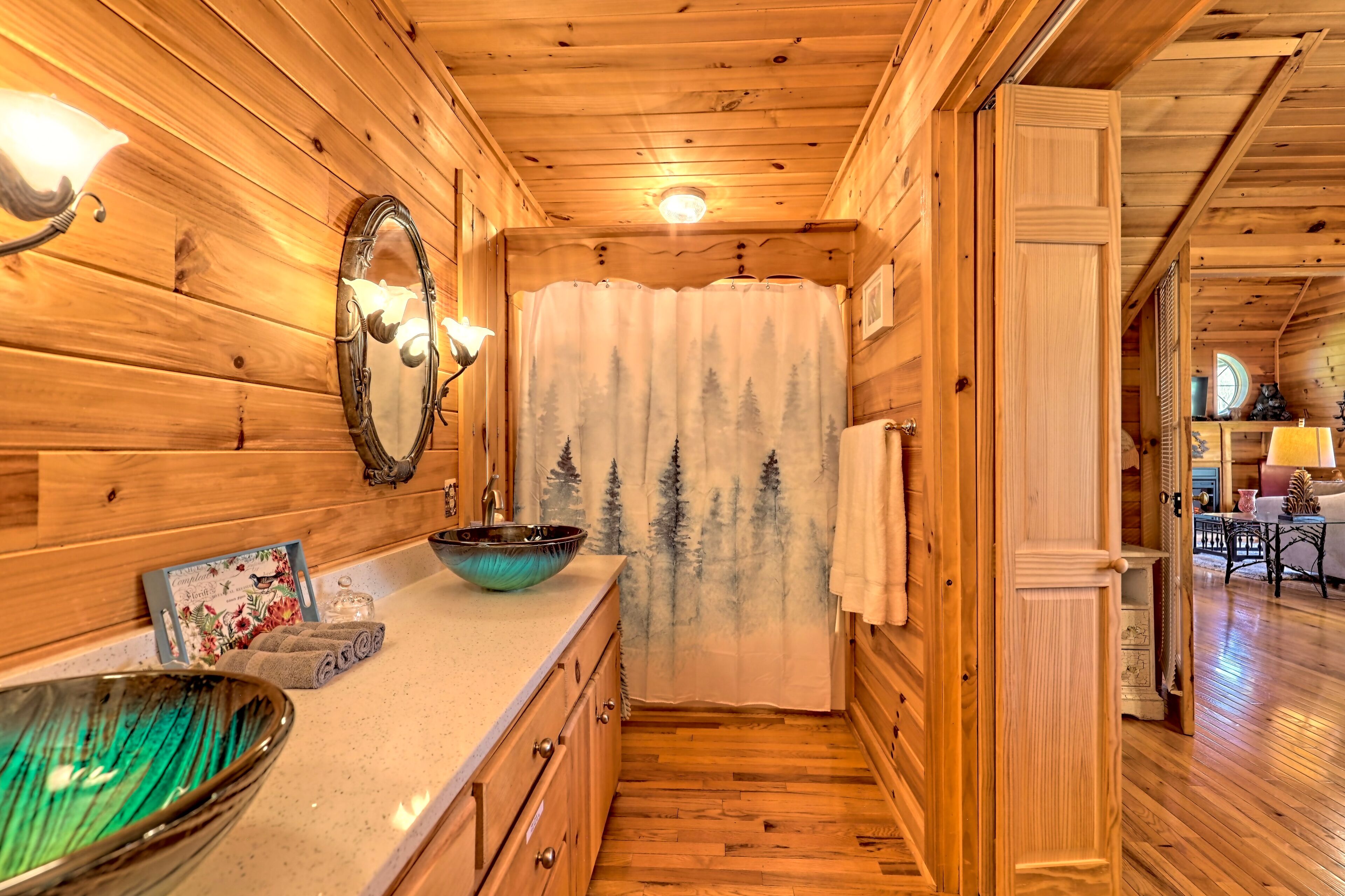 L’intérieur d’un chalet en bois comprend une salle de bain avec un meuble-lavabo en bois, un grand miroir et une fenêtre ornée d’un rideau représentant un paysage forestier.