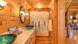 A wooden cabin interior with a bathroom featuring a wooden vanity, a large mirror, and a window with a forest scene curtain.