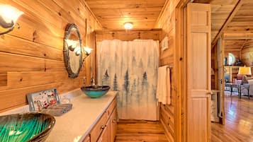 L’intérieur d’un chalet en bois comprend une salle de bain avec un meuble-lavabo en bois, un grand miroir et une fenêtre ornée d’un rideau représentant un paysage forestier.