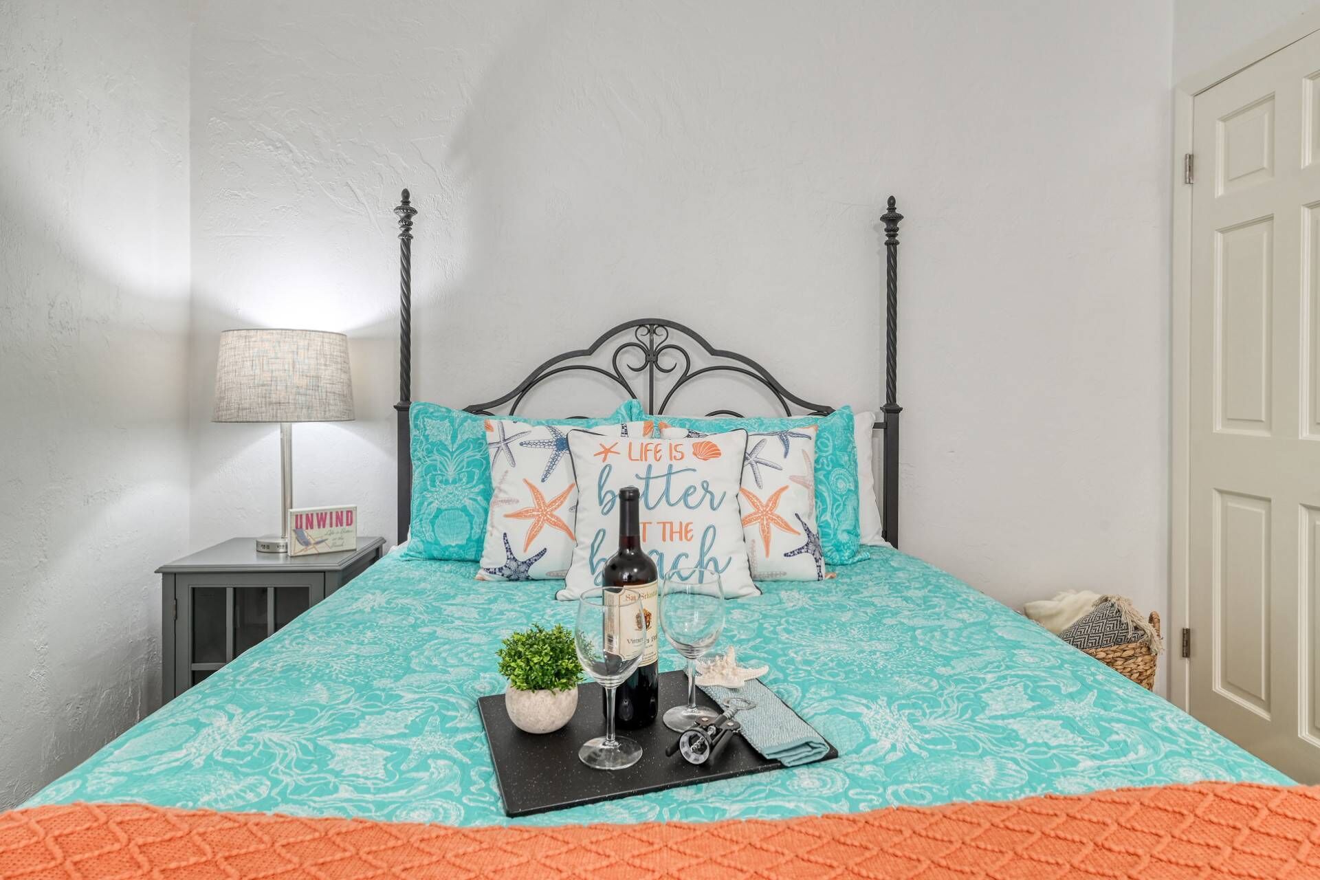 A neatly made bed with a teal and orange bedspread, starfish pillows, a bottle of wine, and a small plant on the bed.