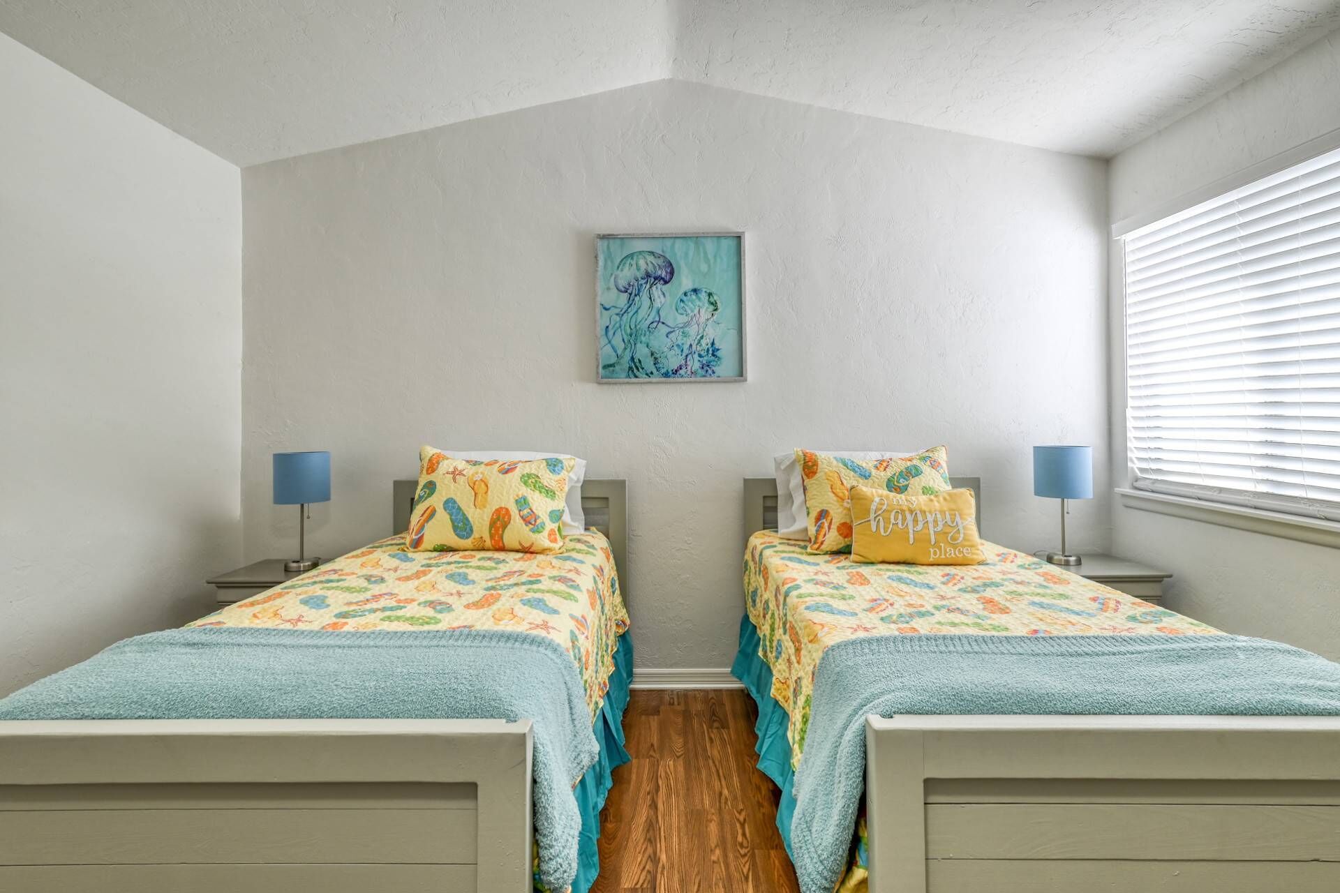 Un dormitorio con dos camas, cada una con un edredón colorido y cojines a juego. Hay una obra de arte enmarcada en la pared sobre las camas, y una ventana con persianas a la derecha.