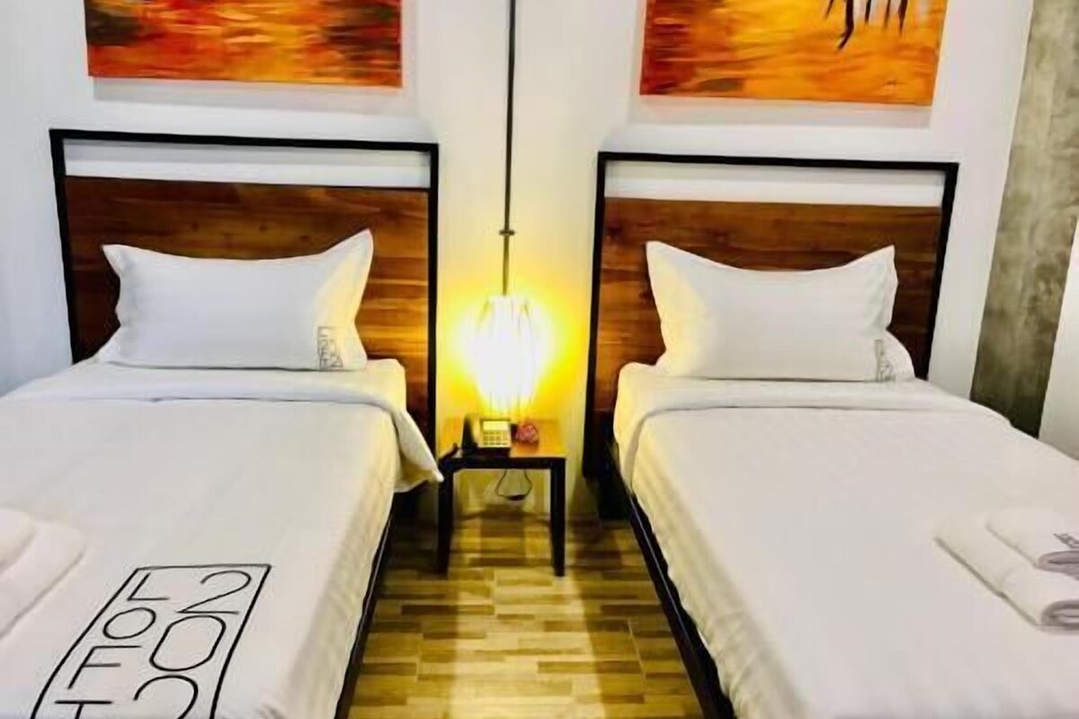 A hotel room with two beds, wooden headboards, white bedding, a wooden floor, and a nightstand with a lamp.