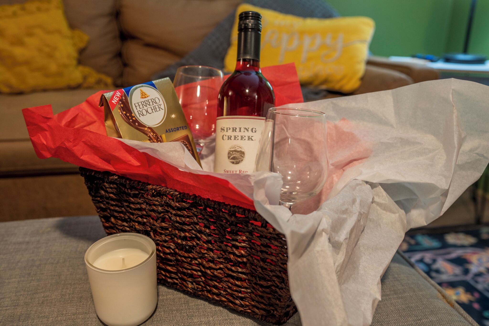 Een rieten mand met een kaars, een fles wijn, een glas en een doos Ferrero Rocher chocolade op een bank.