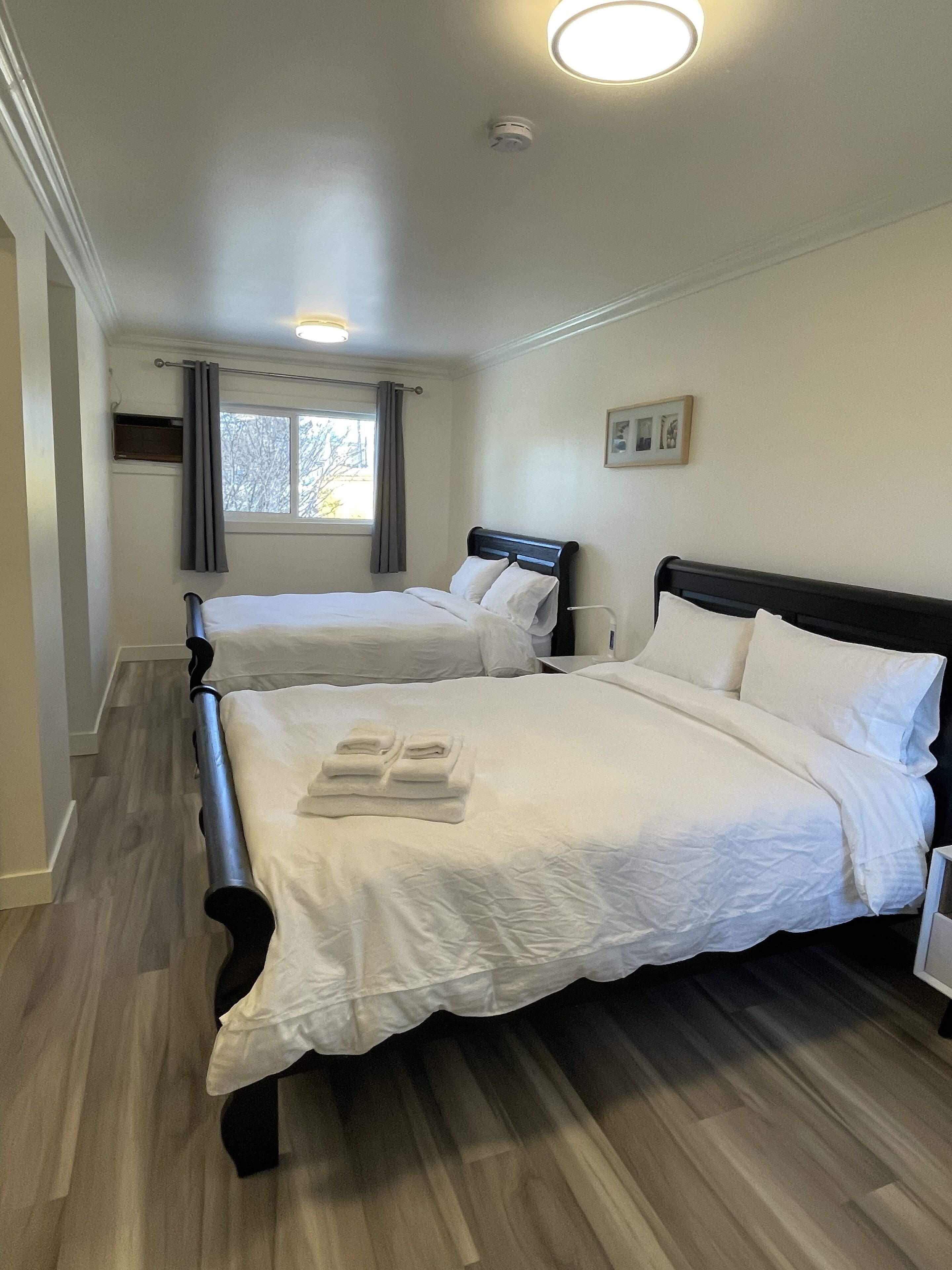 Une chambre d’hôtel avec deux lits, une fenêtre avec des rideaux, un tableau encadré au mur et un luminaire au plafond.