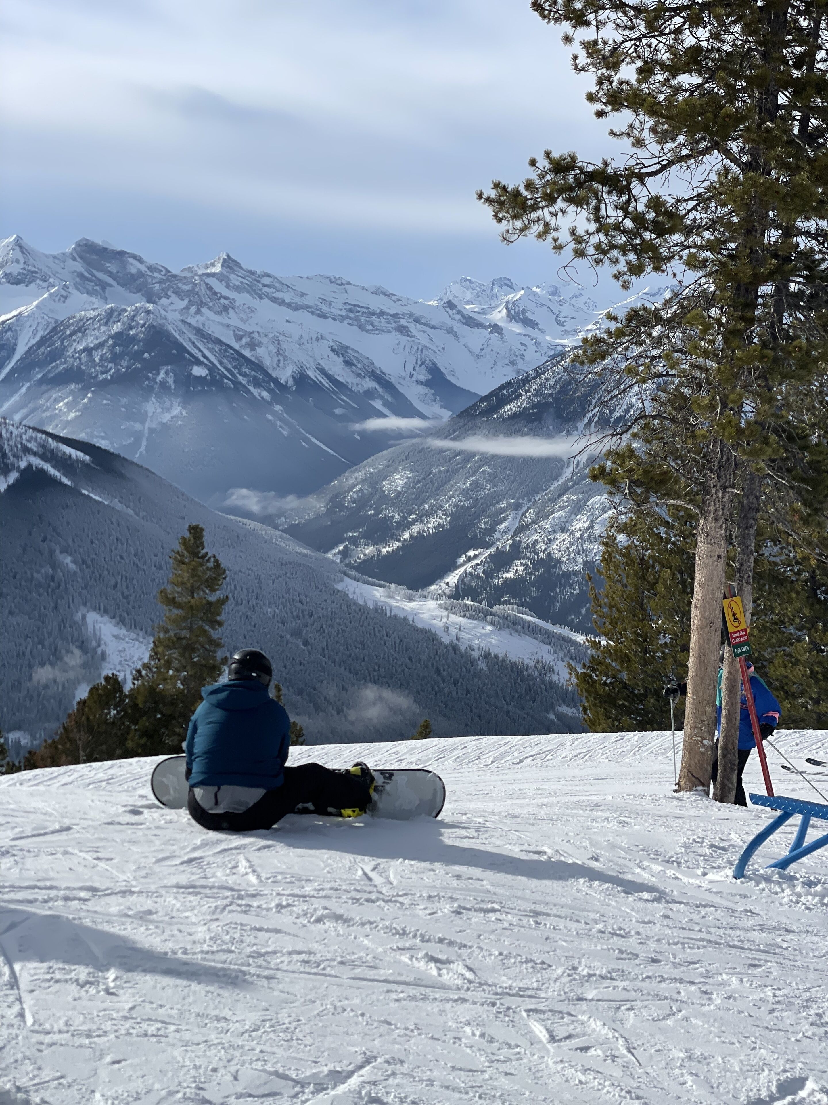 A snowy mountain landscape with a snowboarder sitting on the slope and another person standing nearby.