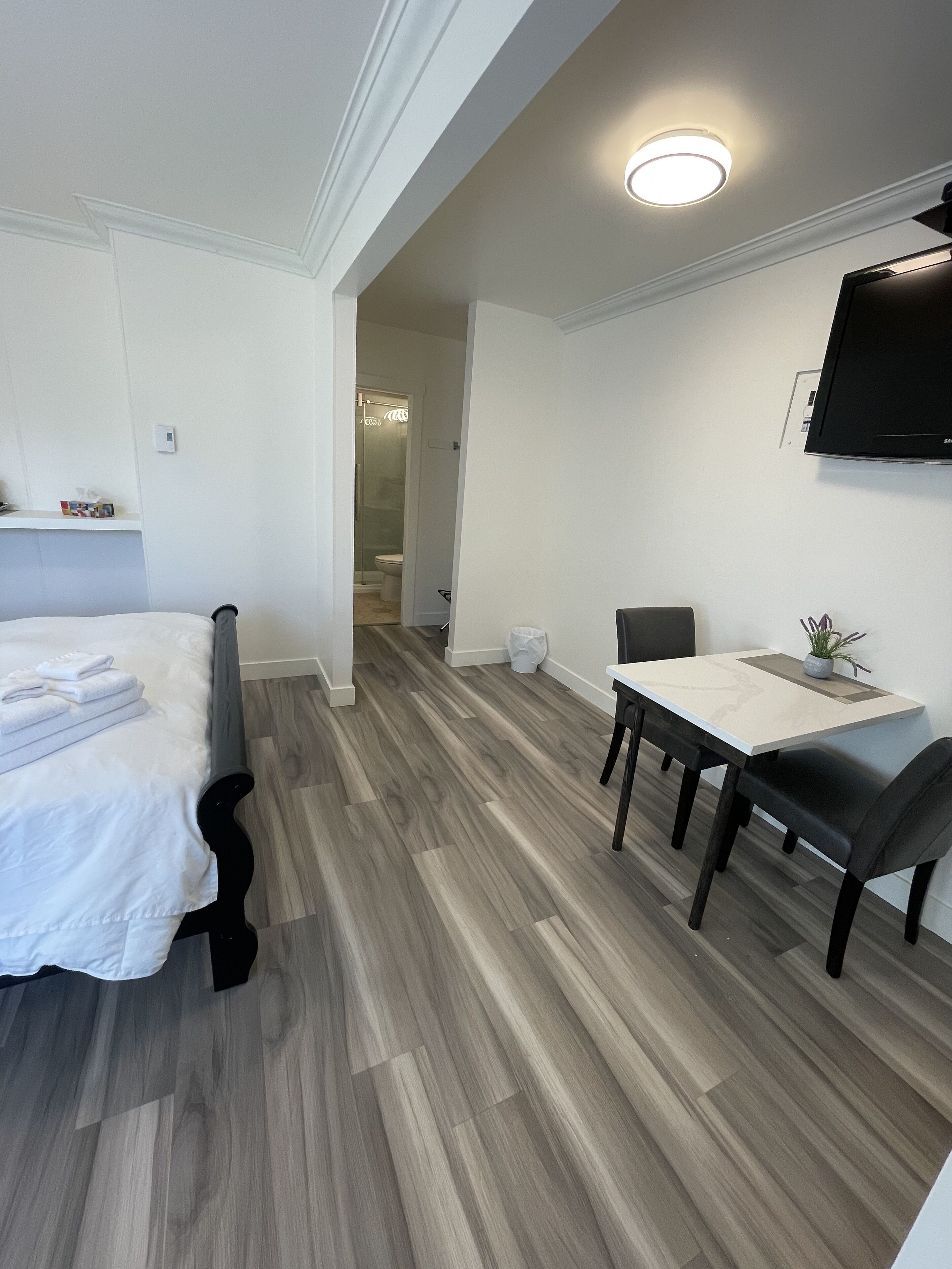 A modern hotel room with a bed, a dining table with chairs, a TV mounted on the wall, and a shower area visible through an open door.