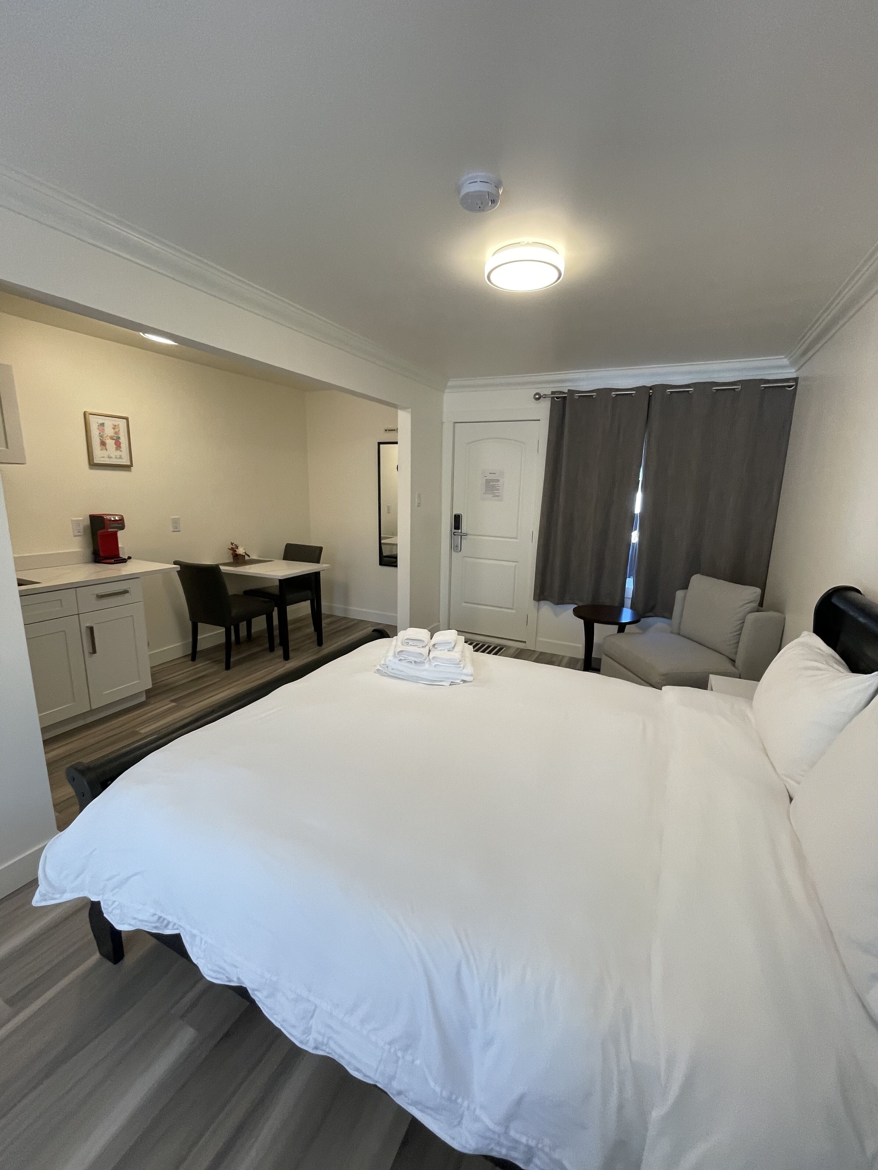 A hotel room with a large bed, a desk with a coffee maker, a chair, a small table, and a door with curtains.