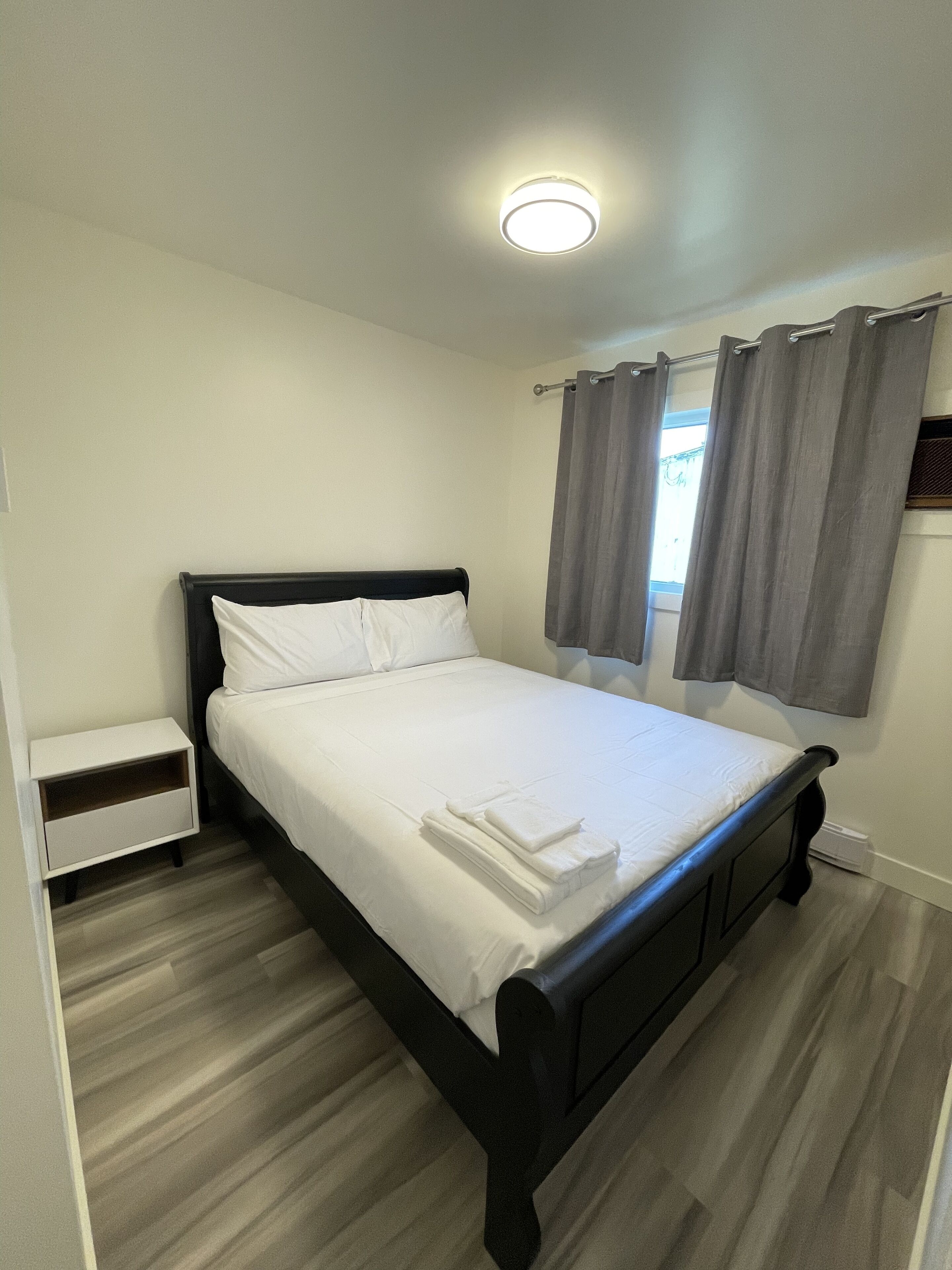 A neatly made bed with white linens and a folded towel, a small bedside table, and a window with grey curtains.