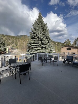 An outdoor patio with tables and chairs, a large evergreen tree, and a view of a mountainous area.