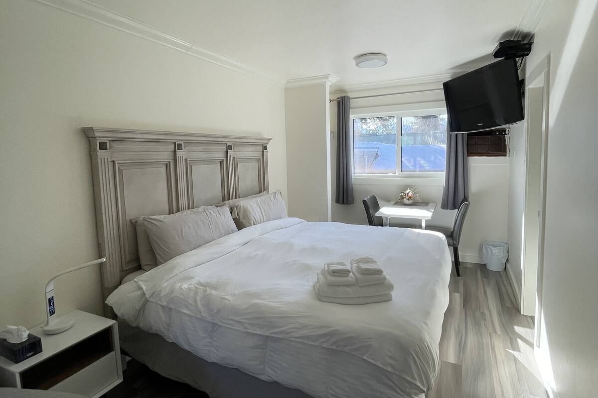 Una habitación de hotel con una cama grande, una mesita de noche con lámpara, un televisor montado en la pared y una ventana con vistas.