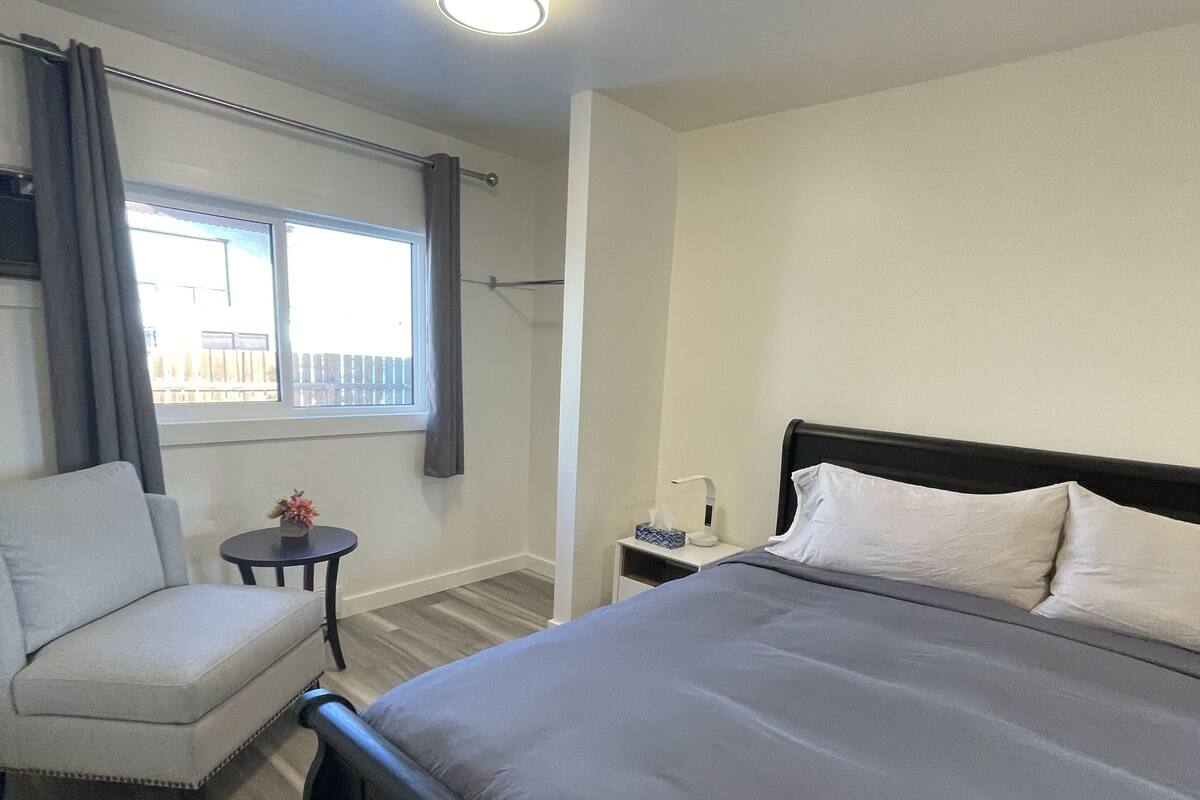 Habitación de hotel con cama, una silla, una mesita, una mesita de noche, ventana con cortinas y una lámpara en el techo.