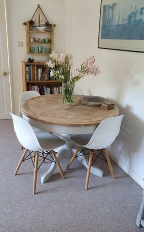 A round wooden dining table with two white chairs, a vase of flowers, and a stack of books on the table.