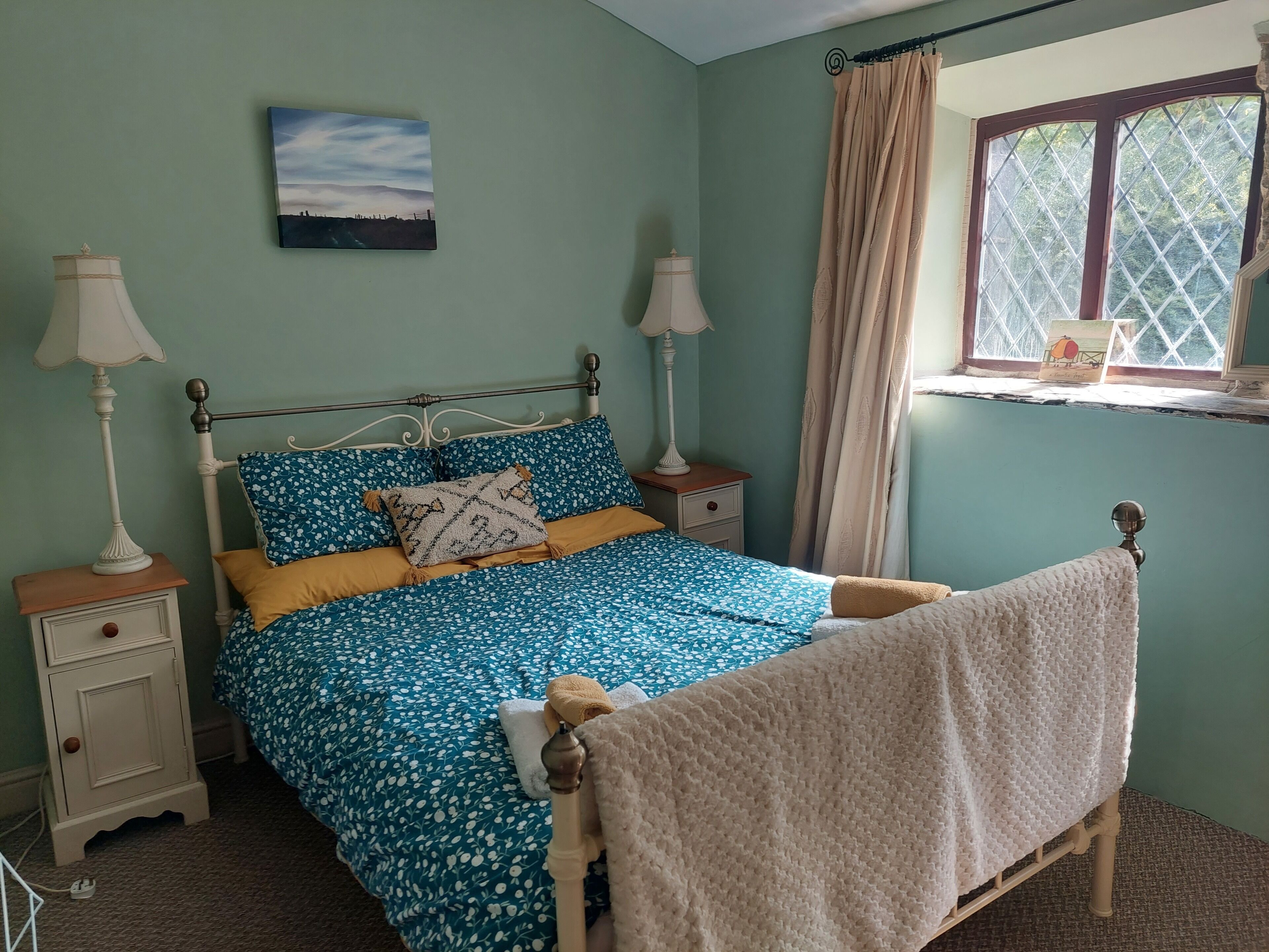 Een slaapkamer met een bed, twee nachtkastjes, een raam met gordijnen, een aan de muur hangend schilderij en een bedlampje.