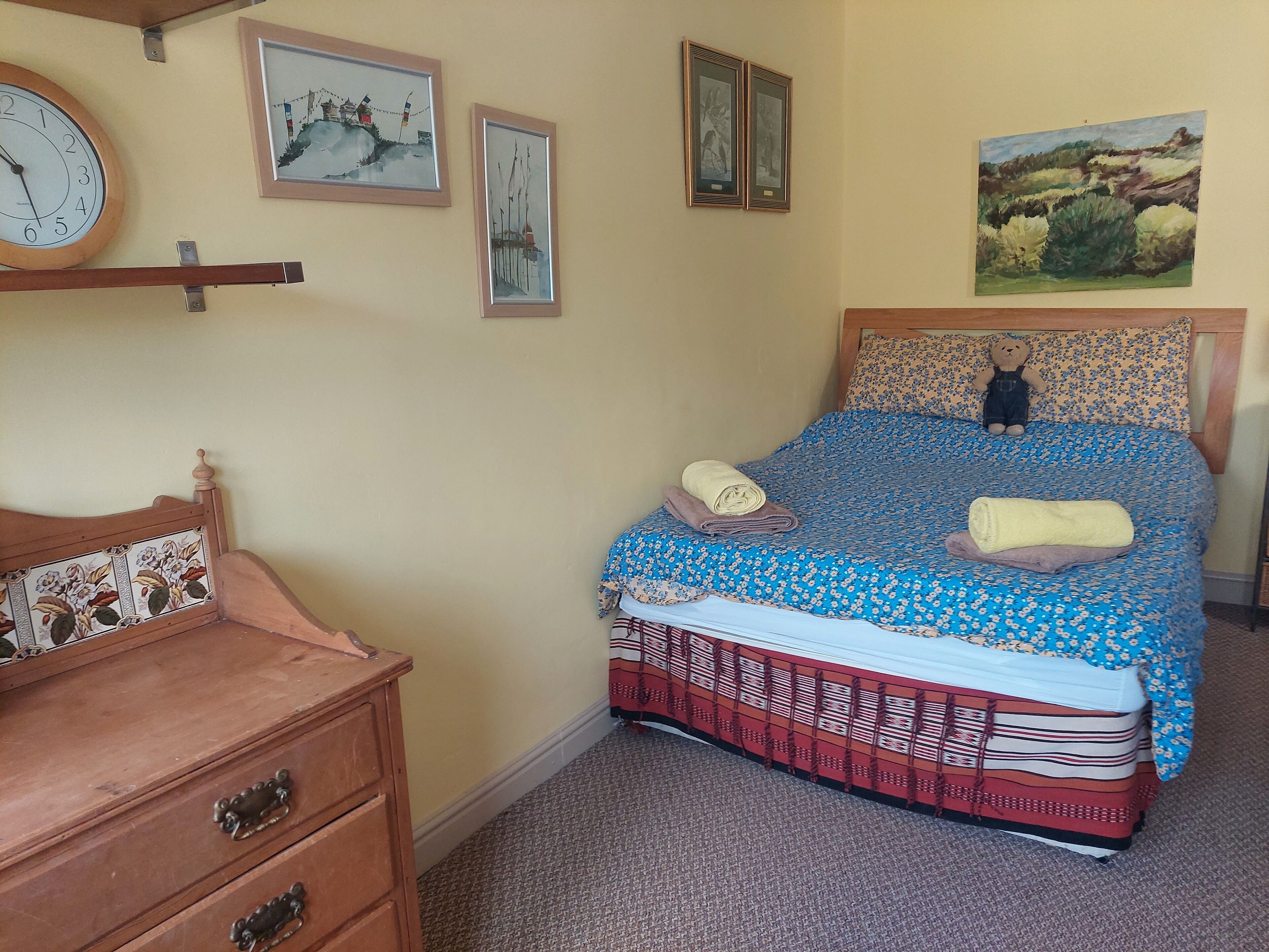 A bedroom with a bed, a wooden dresser, a clock, and wall art.