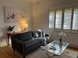 A living room with a black sofa, a glass coffee table, a lamp, and a window with blinds.
