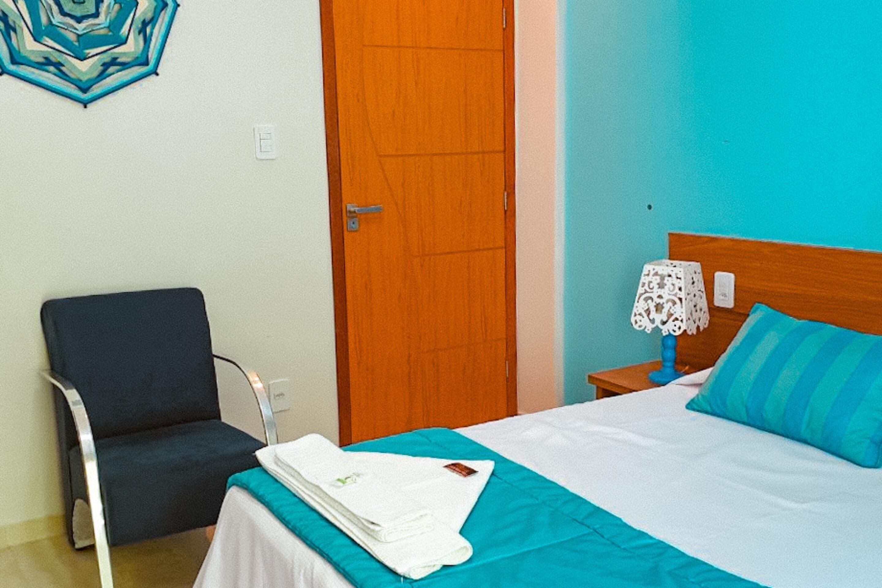 A hotel room with a bed, a chair, a wooden door, and a nightstand with a lamp.