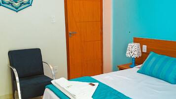 A hotel room with a bed, a chair, a wooden door, and a nightstand with a lamp.