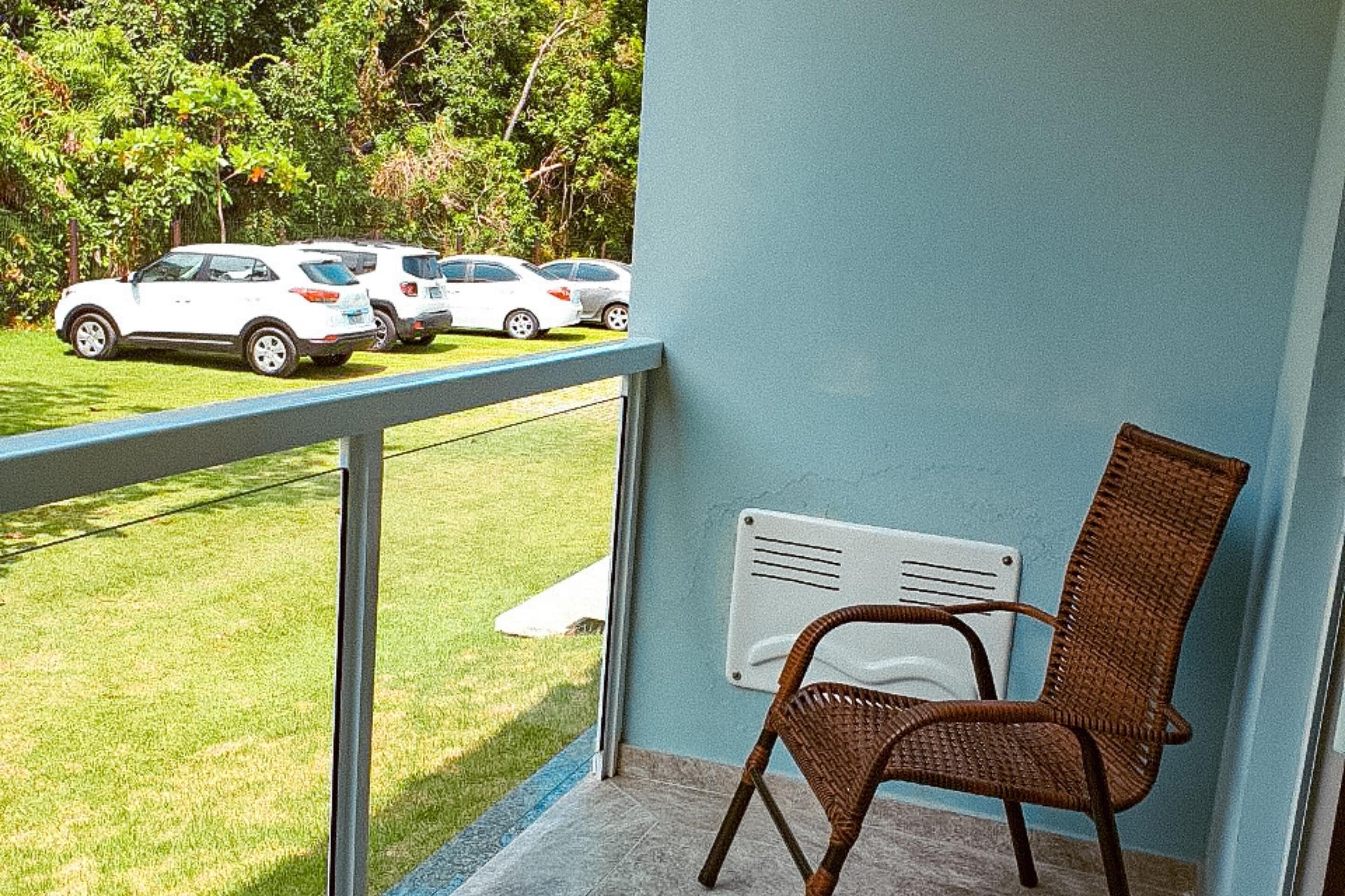 Varanda com cadeira de vime, radiador branco e vista para carros estacionados numa área ajardinada.