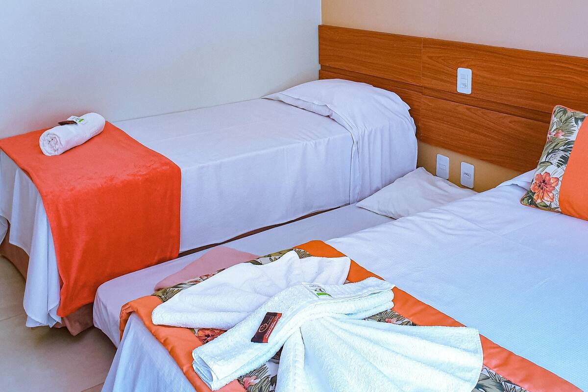 A hotel room with two single beds, each with a white bedspread and an orange blanket. There is a wooden headboard on the right bed and a white wall on the left.