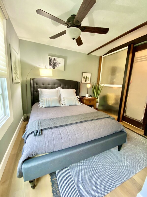 Quarto com uma cama grande, um ventilador de teto, uma mesa de cabeceira com abajur e uma porta de correr de vidro.