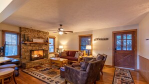 A cozy living room with a stone fireplace, wooden furniture, and a large window with blinds. - Nuttin Better - App Ski Mtn cabin with hot tub, game table (Blowing Rock)
