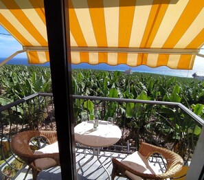 A balcony with a yellow-striped awning, wicker furniture, and a view of a banana plantation.
