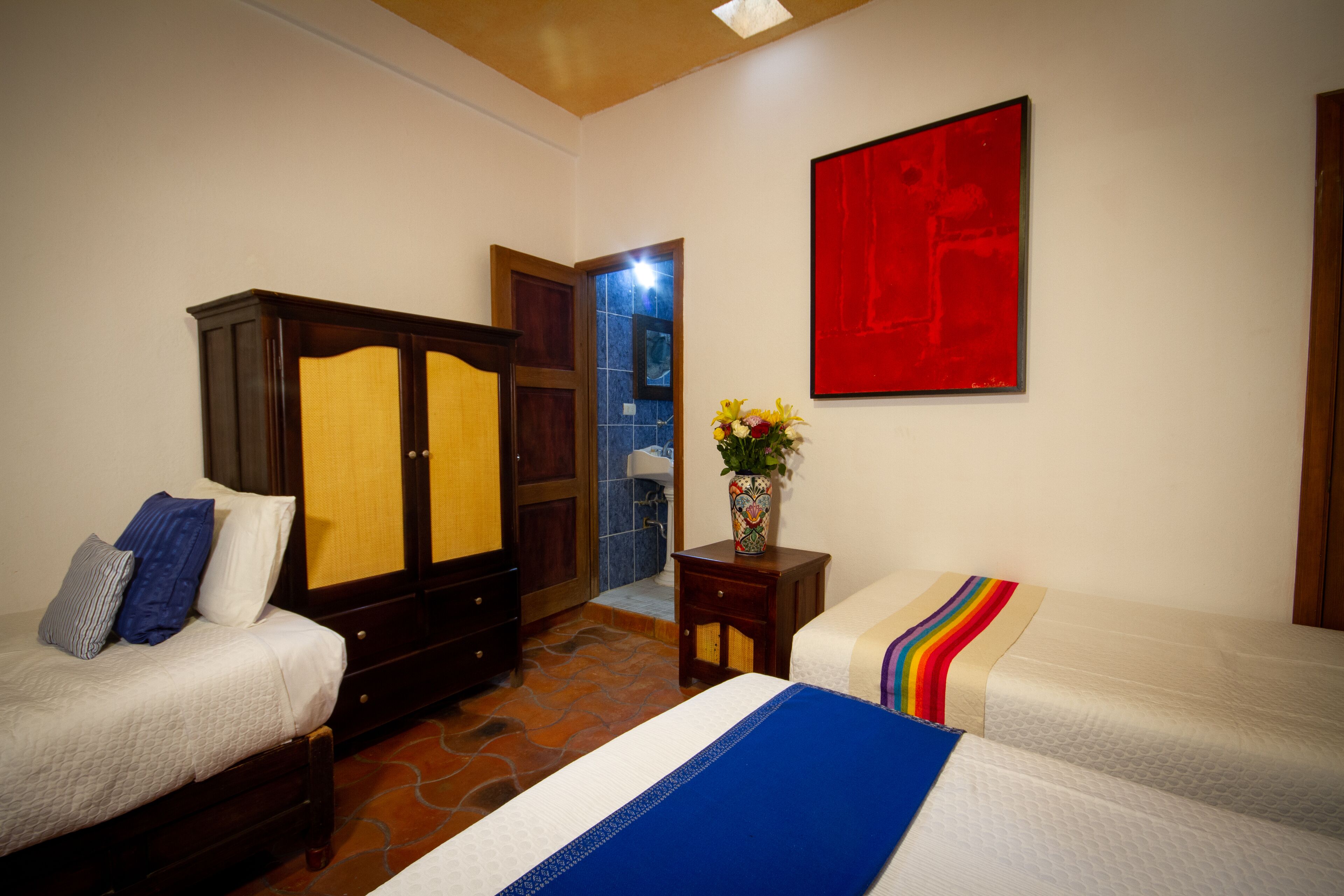 Habitación de hotel con dos camas, un armario, un cuadro abstracto rojo y un florero con flores.