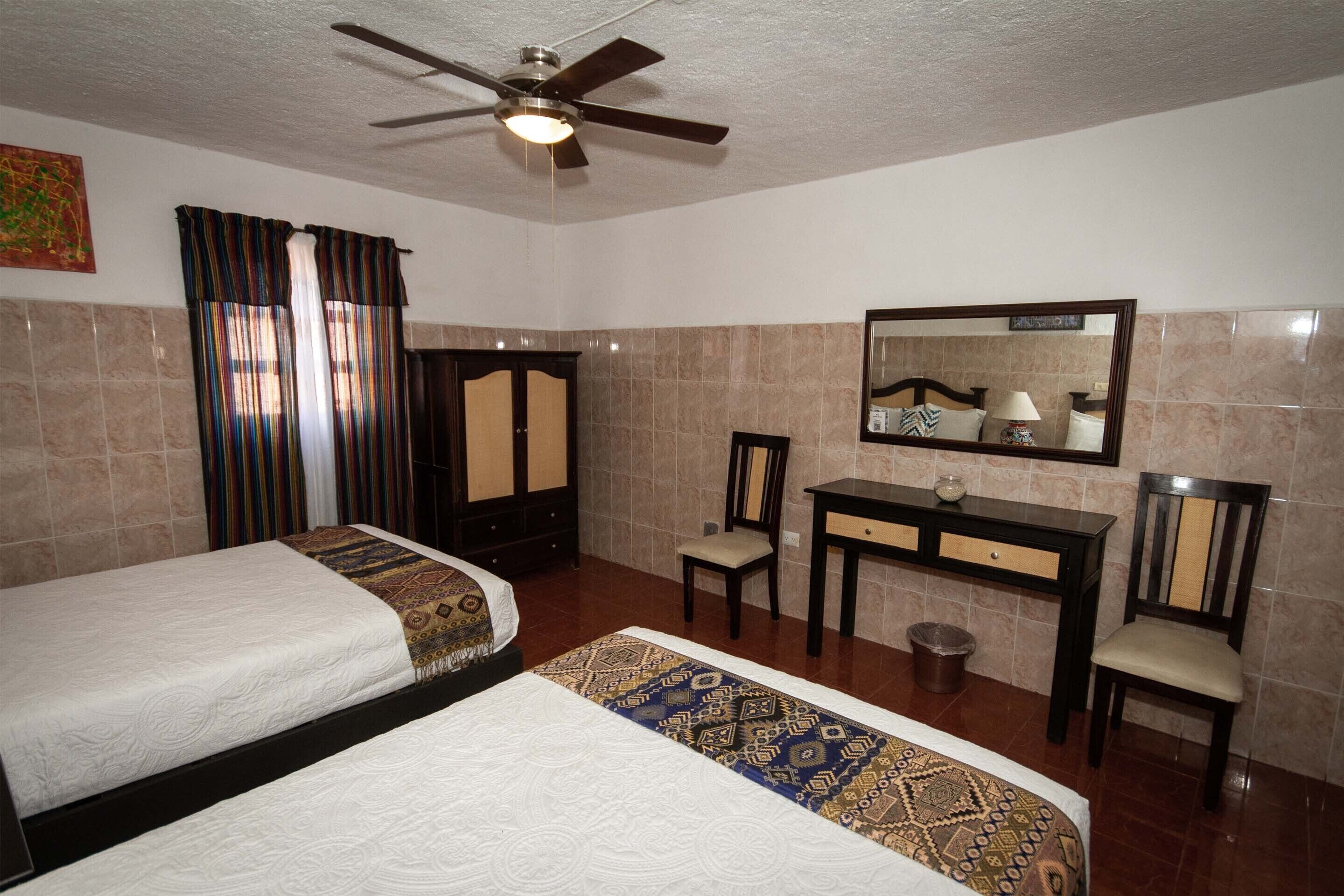A hotel room with two beds, a desk, a chair, a mirror, and a ceiling fan.