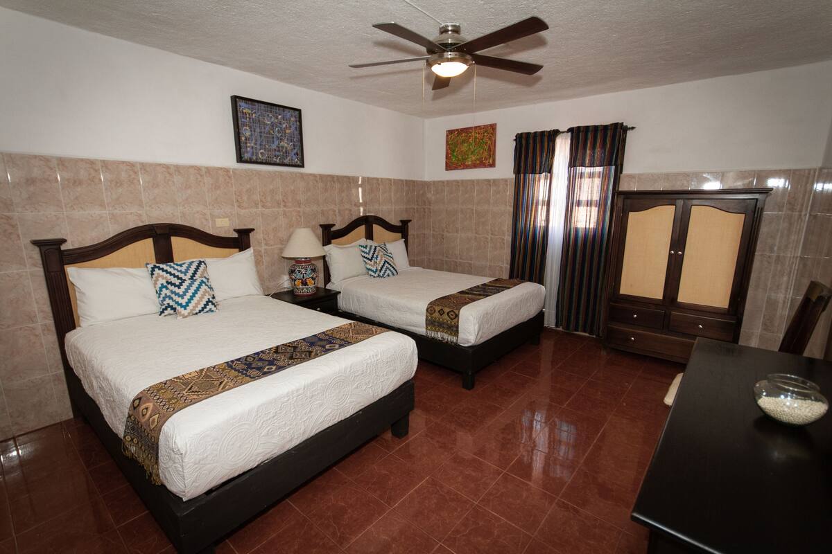 A hotel room with two beds, a ceiling fan, a nightstand with a lamp, a table with a bowl, and a cabinet.