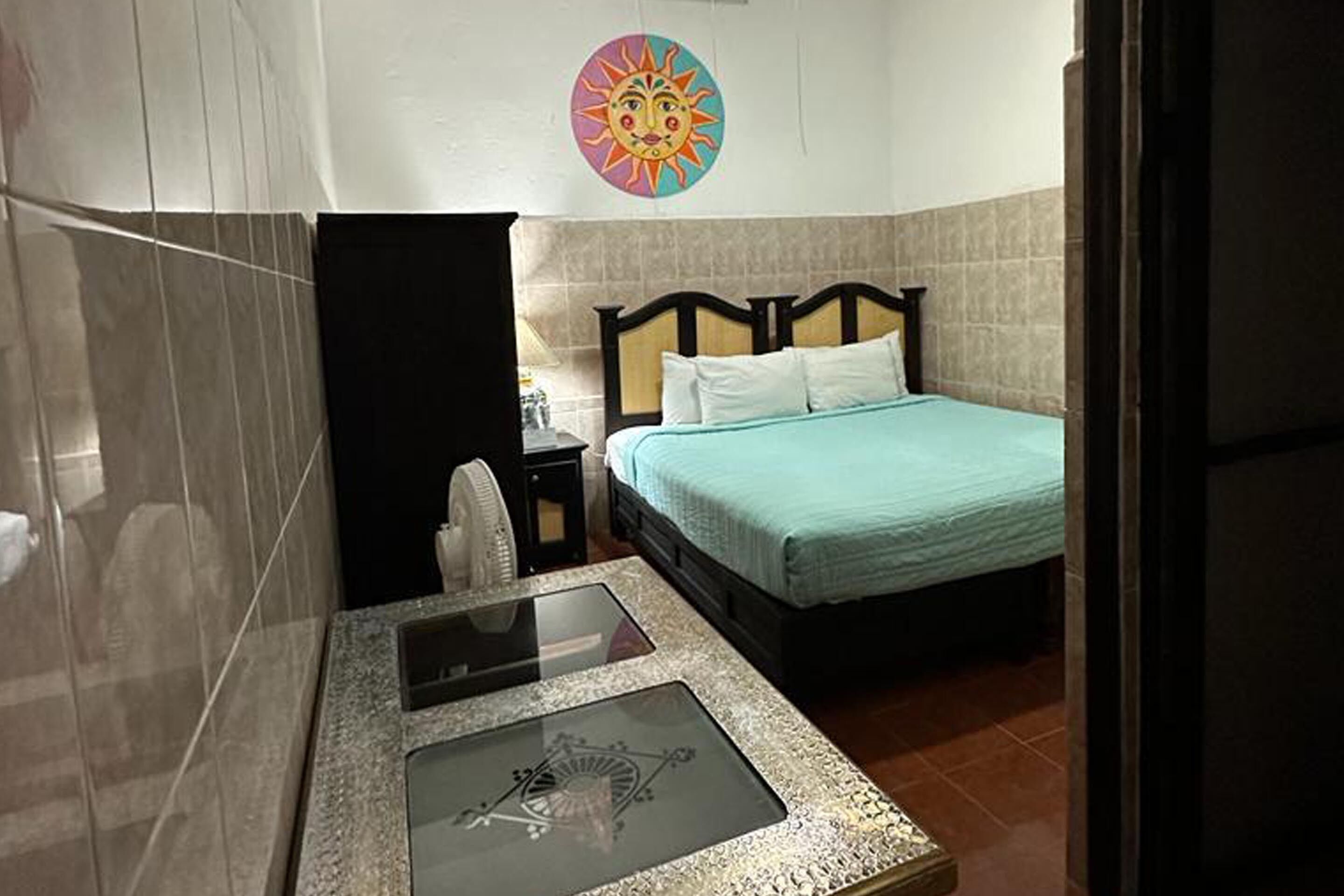 Habitación de hotel con una cama, una mesita con superficie de vidrio, un toallero en la pared y un cuadro decorativo con motivo de sol.