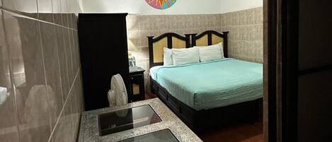 Habitación de hotel con una cama, una mesita con superficie de vidrio, un toallero en la pared y un cuadro decorativo con motivo de sol.
