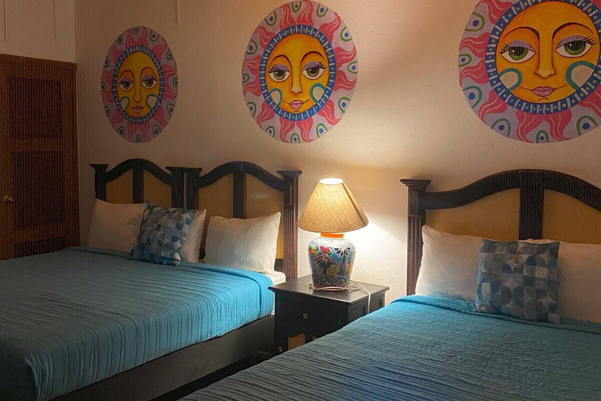 A hotel room with two beds, each with blue bedding and decorative pillows. There is a nightstand with a lamp between the beds. The walls feature circular wall art with face designs.