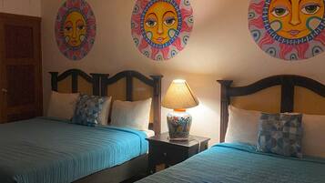 A hotel room with two beds, each with blue bedding and decorative pillows. There is a nightstand with a lamp between the beds. The walls feature circular wall art with face designs.