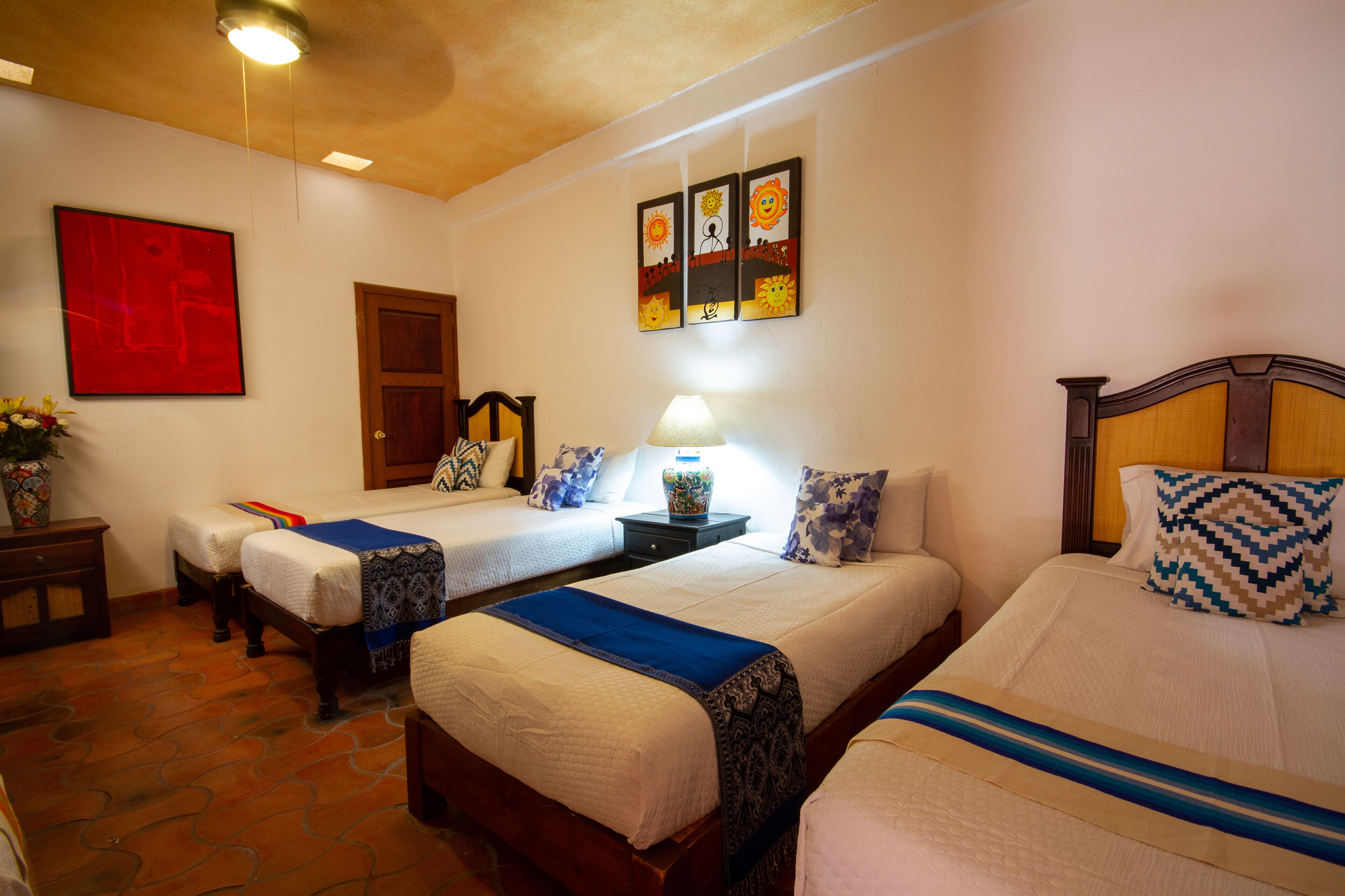 A hotel room with two single beds, a nightstand with a lamp, a red abstract painting, and a wall with three framed drawings.