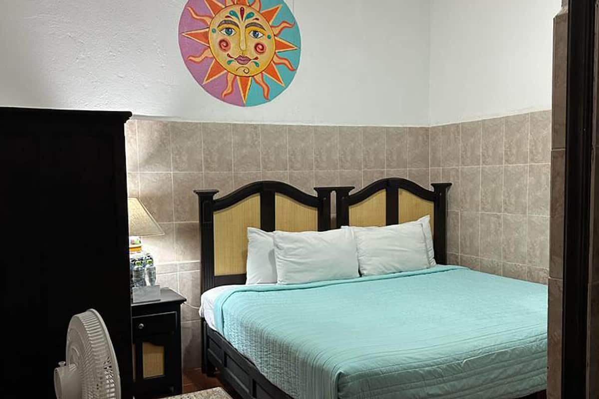 A bedroom with a bed, a nightstand, a wall-mounted fan, and a decorative sun mural.