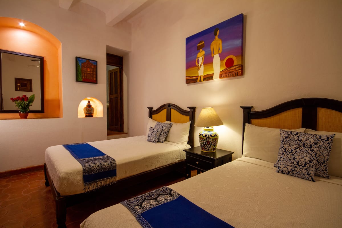 A hotel room with two beds, a nightstand with a lamp, a painting on the wall, and a mirror with a vase of flowers.