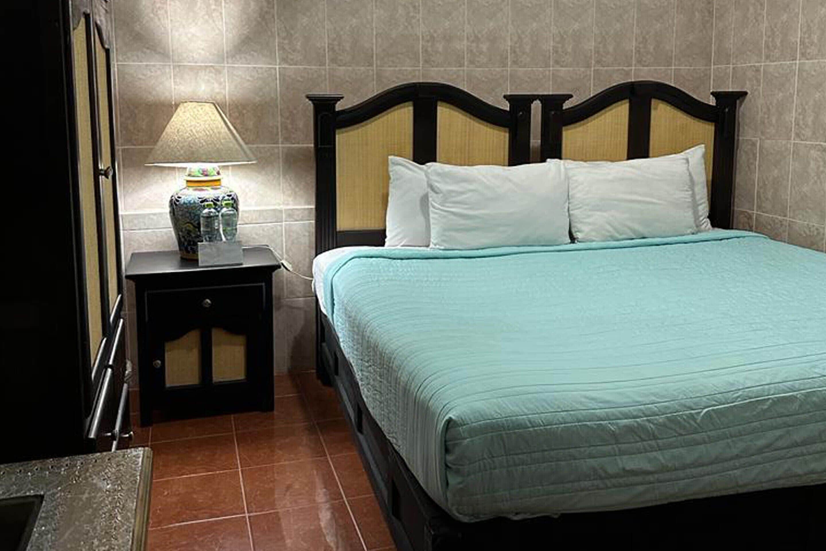 A neatly made bed with a teal bedspread and white pillows, a bedside lamp with a decorative base, and a tiled wall in the background.