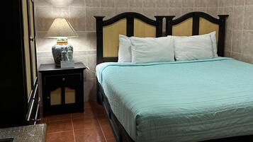 Una cama bien hecha con una colcha color turquesa y almohadas blancas, una lámpara de noche con una base decorativa, y una pared de azulejos al fondo.